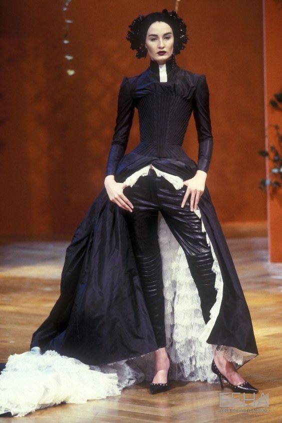 a model is standing on a runway in a striking black highfashion dress. the ensemble features a corsetlike bodice, long sleeves, and a dramatic skirt with layers of white lace. the model is also wearing tight black leggings and pointed black heels. she has a dark, stylized headpiece. the overall look is theatrical and highend.