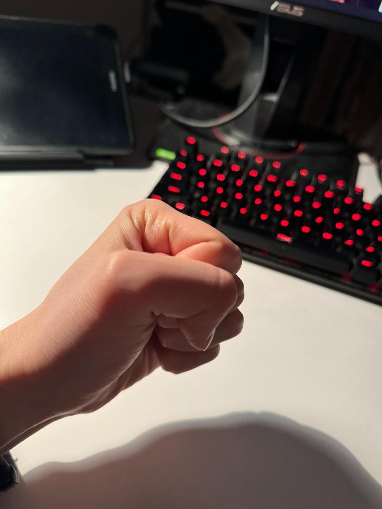 the image is a firstperson perspective of a clenched fist hovering over a white desk. the hand is pale, and the knuckles are prominently displayed. in the background, a black keyboard with glowing red keys sits on the desk. the keyboard suggests a gaming or computer setup. the overall lighting is dim and artificial. the image evokes feelings of anger, frustration, or rage, potentially related to gaming or work.