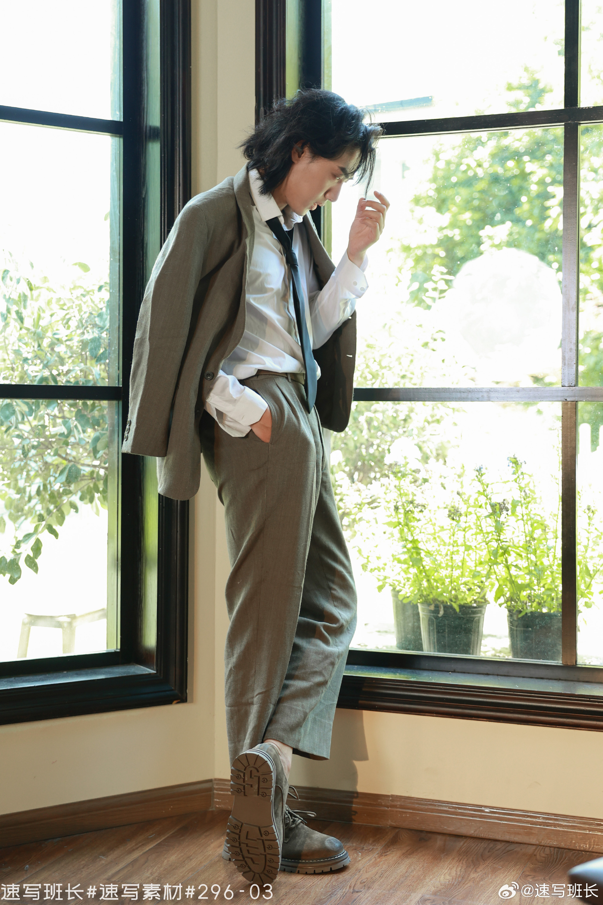 a man in a brown suit and white shirt is standing by a window, looking down with his hand on his head. he is wearing black boots and a black tie. there are plants visible outside the window. the image is taken from eye level.