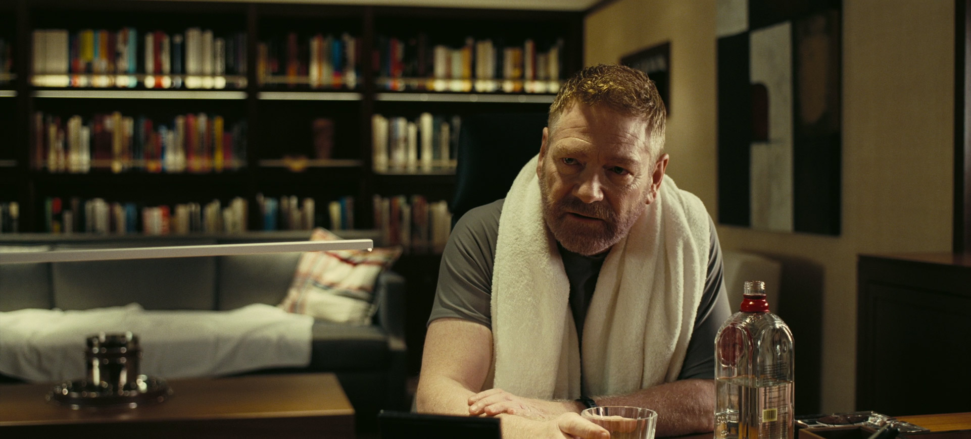 a man with salt and pepper hair is sitting at a table in a dark room, looking intently at something offcamera. he has a white towel wrapped around his neck and a glass of something clear in front of him. there is a bottle of dark liquid behind the glass. to the man's left is a dark sofa and a dark bookshelf filled with books.  the lighting in the room is dim and there are shadows.