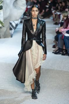 a female model walks the runway, dressed in a long black leather jacket, a white ruffled gown, and black boots. the model is accessorized with a silver necklace and has long dark hair. the model is captured midstride. the model is walking toward the camera, but she is not looking at the camera. the runway is lined with people, and the model is surrounded by them. the people in the background are not visible, but their silhouettes are visible.