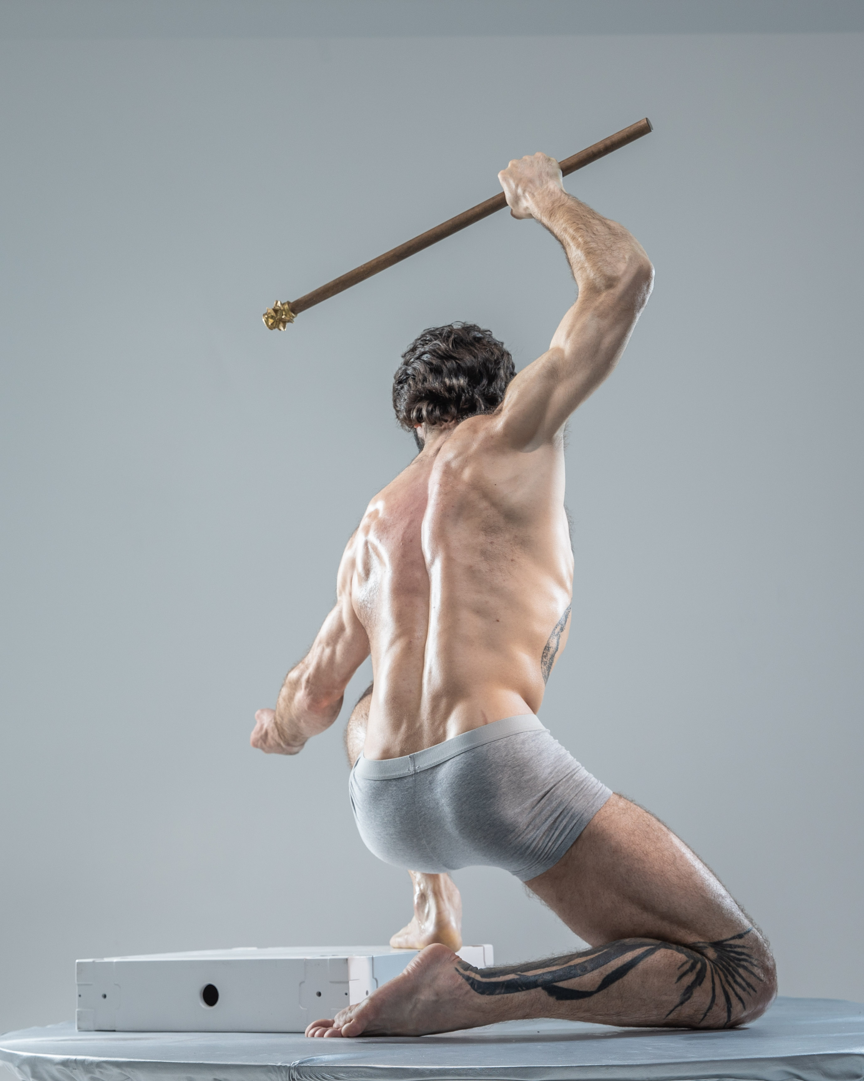 the image showcases a muscular man in a dynamic pose. he's holding a wooden staff above his head with his right hand, seemingly about to strike. he's wearing only gray underwear, revealing a toned physique and a tribal tattoo on his left leg.  the setting appears to be a studio with soft, diffused lighting, highlighting the man's musculature and the tension in his pose. the low angle perspective emphasizes his strength and power.