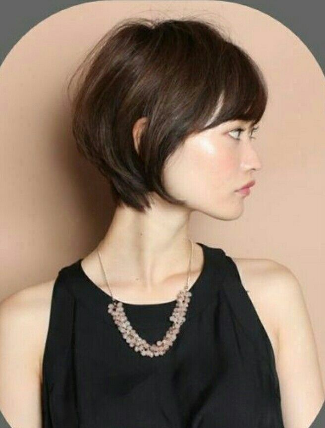 a young woman with short, black hair and bangs is wearing a black dress and a delicate silver necklace. she is smiling slightly as she looks off to the side. the background is a light brown color.