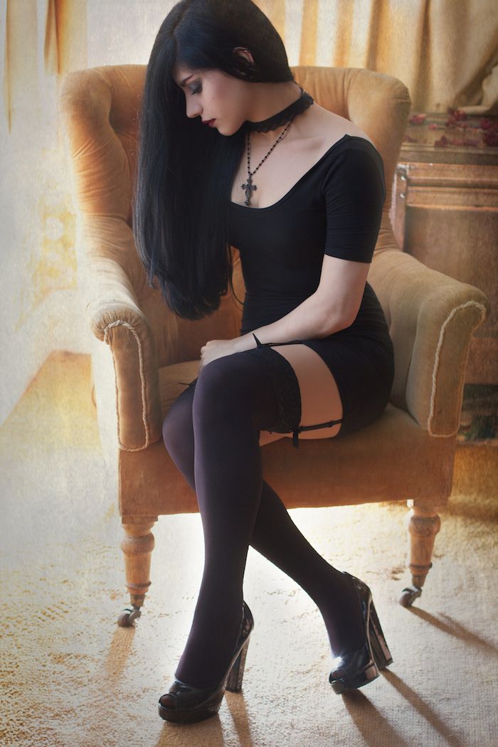 a young woman with long, dark hair and pale skin is seated in a vintage armchair. she wears a simple black dress, black thighhigh stockings, and black heels. a black choker with a silver cross pendant adorns her neck. her head is bowed, and her expression is melancholic. the environment suggests a vintage interior, with warm, soft lighting casting shadows in the background.