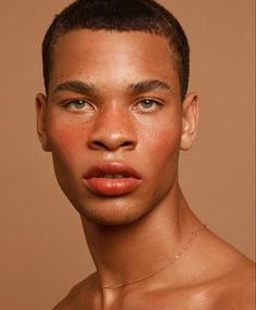 a closeup headshot of a young male model with short hair, looking directly at the camera. he has brown eyes, full lips, and freckles across his nose and cheeks. his skin is smooth and has a warm, peachy undertone. the lighting is soft and creates a subtle highlight on his forehead and cheekbones. he is wearing a thin gold chain around his neck. the background is a solid tan color.