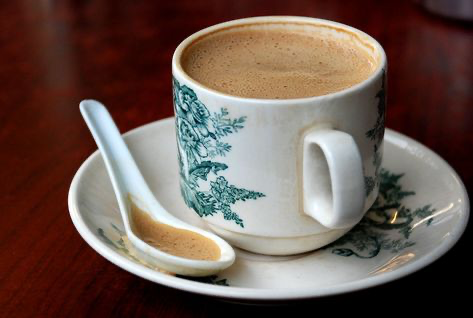 a closeup shot of a cup of coffee on a saucer with a small white spoon. the cup and saucer are decorated with a floral pattern. the coffee is dark brown and appears to be hot. the table is made of brown wood.