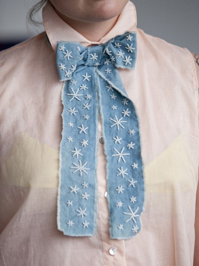 the image shows a closeup of a woman wearing a unique, handmade bow tie. the bow tie is made from light blue denim with raw edges and is decorated with white pearl embroidery in a snowflake pattern. the bow tie is worn over a pale pink, sleeveless cotton shirt. the overall style is bohochic and the image emphasizes the craftsmanship and detail of the handcrafted accessory.  the woman's face is partially visible, but the focus is primarily on the bow tie and its intricate details.