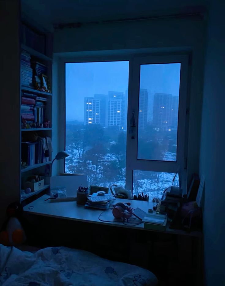 the image shows a cozy bedroom illuminated by the blue light of a winter evening. a large window overlooks a cityscape, revealing snowdusted buildings and a twilight sky. a desk sits in front of the window, cluttered with books, stationery, and headphones. the overall atmosphere is tranquil and contemplative.