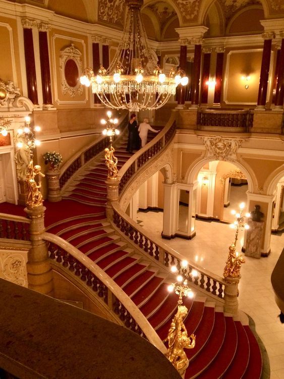 the image showcases a magnificent, grand staircase in a palatial building, possibly an opera house or theatre.  the staircase is carpeted in rich red and features intricate, ornate gold balustrades and golden statues flanking its curves. a large, glittering crystal chandelier hangs above, illuminating the space. two women in elegant evening gowns are visible ascending the stairs. the overall aesthetic is opulent and lavish, showcasing classical architectural details including marble finishes and golden accents. the warm lighting enhances the luxurious atmosphere of the scene.