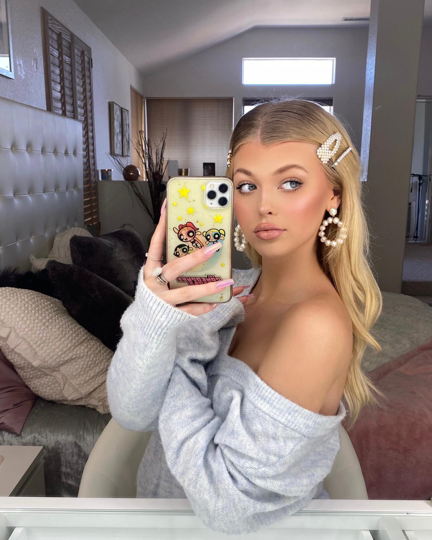 a woman with blonde hair is taking a selfie in a bedroom. she is wearing pearl earrings, hair clips, and an offshoulder sweater. she is holding a phone with a powerpuff girls phone case. the bedroom has a bed with pillows and a mirror.