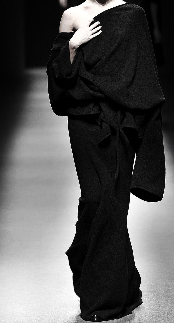 a model walks the runway wearing a long black draping dress. the dress has long sleeves that are draped over the shoulders. the model is striking and the dress is very stylish.  the image is black and white.  the image looks to be taken from a runway show.