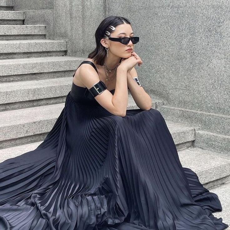 a woman is sitting on concrete stairs, wearing a long, black pleated dress. she is also wearing sunglasses, hair clips, jewelry, and arm cuffs. she has a serious expression on her face and her hands are resting beneath her chin. the background consists of the stairs and a concrete wall.