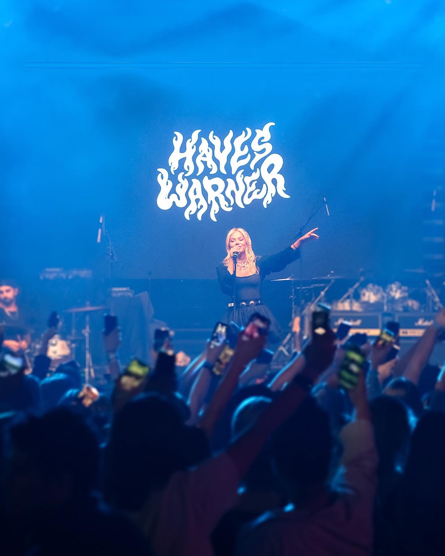 a photo of a concert featuring hayes warner performing on stage. the stage is illuminated with blue lighting and has a large screen behind her displaying the name "hayes warner" in a stylized font. warner, a blonde woman dressed in a dark outfit, is singing into a microphone with one hand raised. in front of the stage, a large crowd of people is holding up their smartphones to record the performance. there are visible band members and musical equipment on the stage.