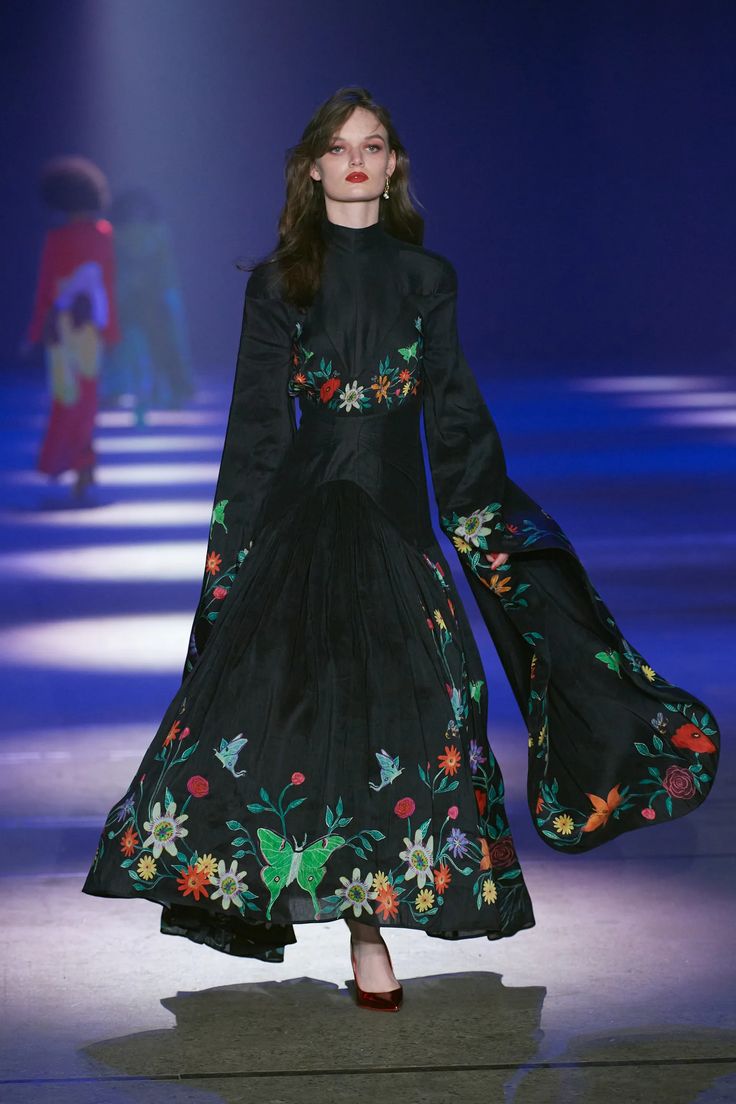 a fashion model walks down a runway wearing a longsleeved, floorlength black dress with intricate floral embroidery. the dress features a high neckline, a fitted bodice, and a full, pleated skirt. the sleeves are wide and flowing. the model has red lipstick and wears red heels. the background is a bluelit stage with other models in the distance.