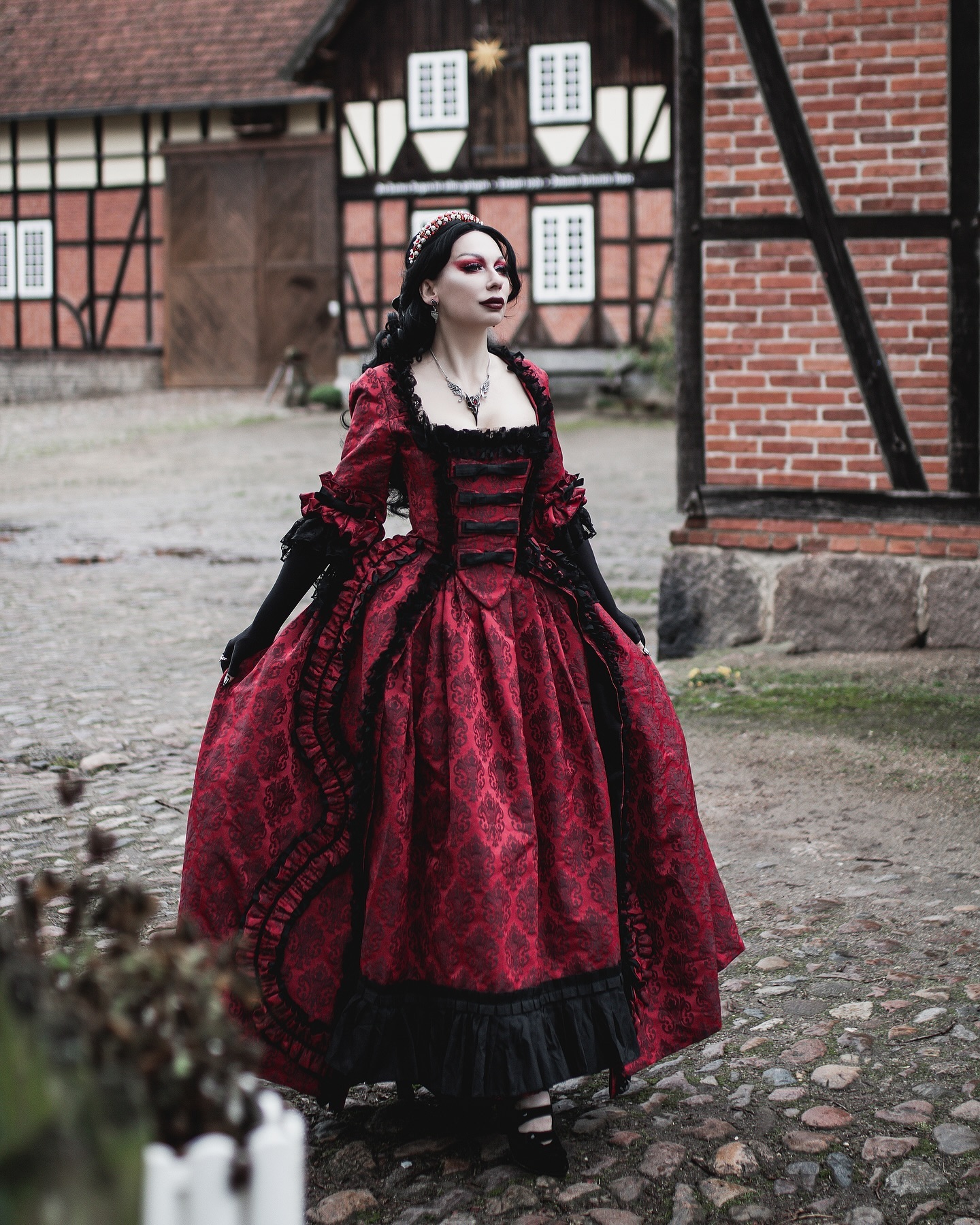 a woman in a dark red and black gothic victorianstyle dress stands outdoors in front of an old building. the dress is long and ornate, with black lace and a corsetlike bodice.  the setting is a cobblestone courtyard with a halftimbered building in the background. the woman's makeup is dark and dramatic, enhancing the overall gothic aesthetic.  she wears a crown and necklace. the overall mood is dark, mysterious and romantic.