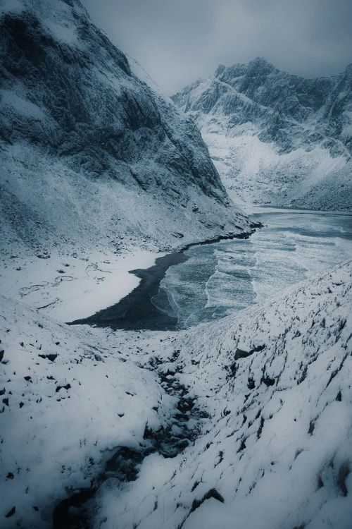 this image shows a snowcovered valley between mountains. a frozen lake sits at the end of the valley. a stream flows from the valley floor. the mountains and ground are completely covered in snow.