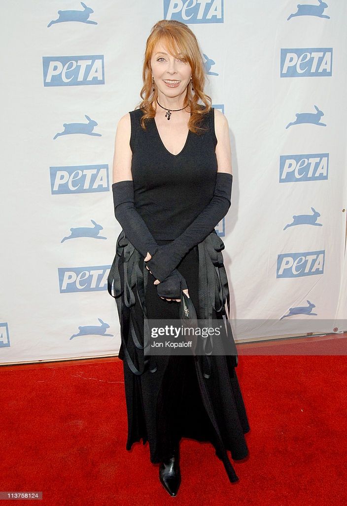 a woman with long red hair is posing on a red carpet in a black dress, long gloves, and black heels. she is looking at the camera, and the background is a white wall with the peta logo printed on it.