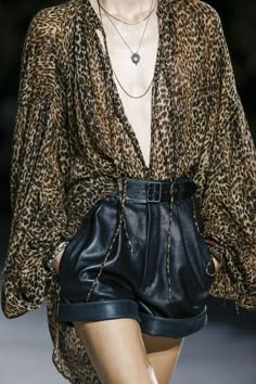 a fashion model on a runway is wearing a leopard print blouse and black leather shorts. she is accessorized with a silver necklace and a belt with a silver buckle. the model's hair is styled in a sleek, straight fashion. the outfit is chic, glamorous, and trendy.