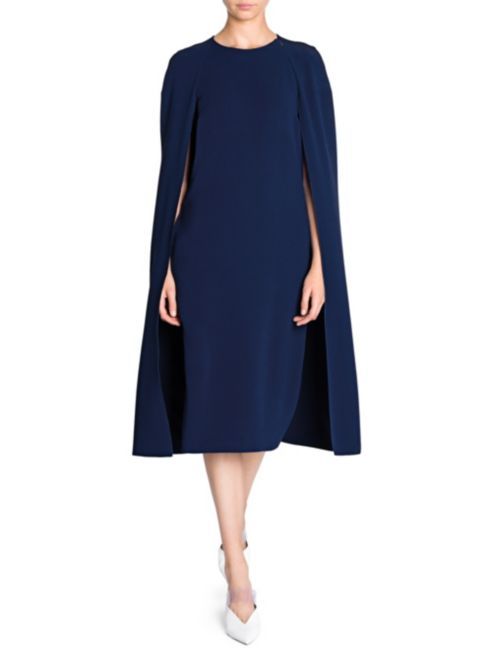 a fulllength studio shot of a woman modeling a navy blue cape dress. the dress has a midi length, aline silhouette, and a simple, collared neckline.  the cape is long, flowing, and seamlessly integrated with the dress.  the model is wearing white pointedtoe shoes. the overall style is formal and elegant. the background is plain white, drawing all attention to the clothing.