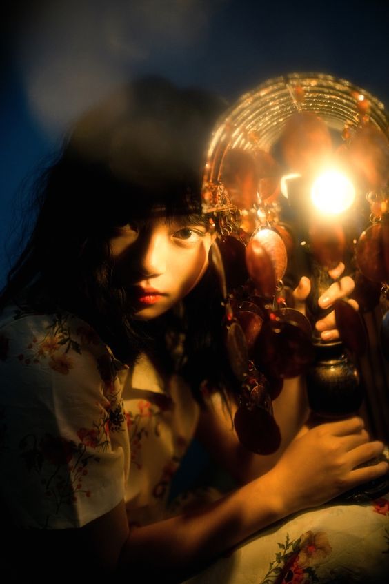 a young girl sits in the dark, her face illuminated by the warm glow of an ornate lamp.  the lamp features a large, round, beaded shade and a lit bulb, casting a soft, golden light that highlights her features. the rest of the image is shrouded in shadow, creating a sense of mystery and intimacy.