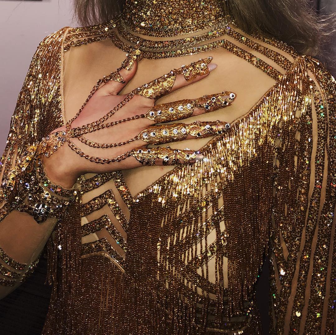 a closeup shot showcases a person adorned in a gold, heavily embellished outfit with matching hand jewelry. the ensemble features a highneck design, long sleeves, and intricate sequin and bead work. the fringe detailing adds texture and movement to the garment. the hand jewelry includes rings on each finger connected by chains to a wrist piece, all in a coordinating gold tone. the sheer fabric elements provide glimpses of skin, adding a touch of allure to the opulent and sparkly aesthetic.