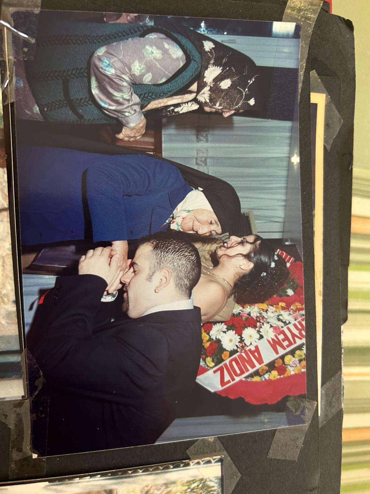 the photograph appears to be from a wedding, possibly a turkish wedding, judging by the attire and the floral arrangement which has a sash with writing on it.  the central focus is a young man in a suit formally kissing the hand of an elderly woman dressed in a blue coat and headscarf. another older woman, wearing a patterned headscarf and vest, stands nearby.  a bride in a lightcolored dress is visible in the background.  the overall scene depicts a moment of respect and tradition, likely involving a blessing or a formal greeting during the wedding ceremony. the photo is a print from a photo album, showing some wear and tear around the edges.