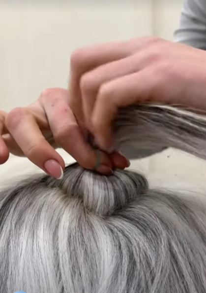 a closeup shot of a woman with silver hair creating a hair bun. her hands are holding the hair together with a small blue hair tie. the hair is neatly wrapped into a bun at the back of her head.