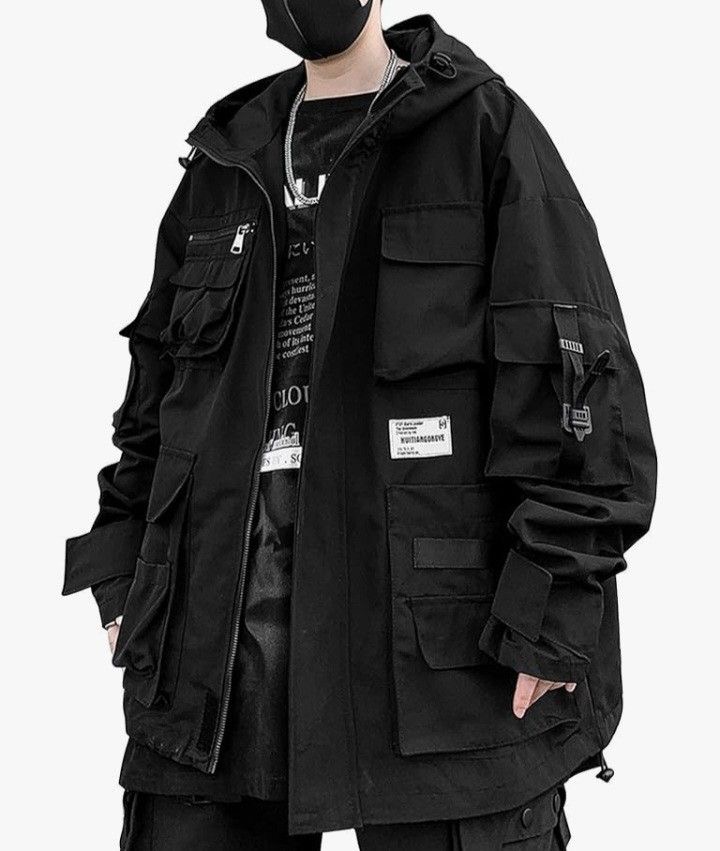 a man wearing a black utility jacket with a hood, multiple pockets, and straps. the jacket is zipped up and the model is looking down. the jacket is a streetwear style.