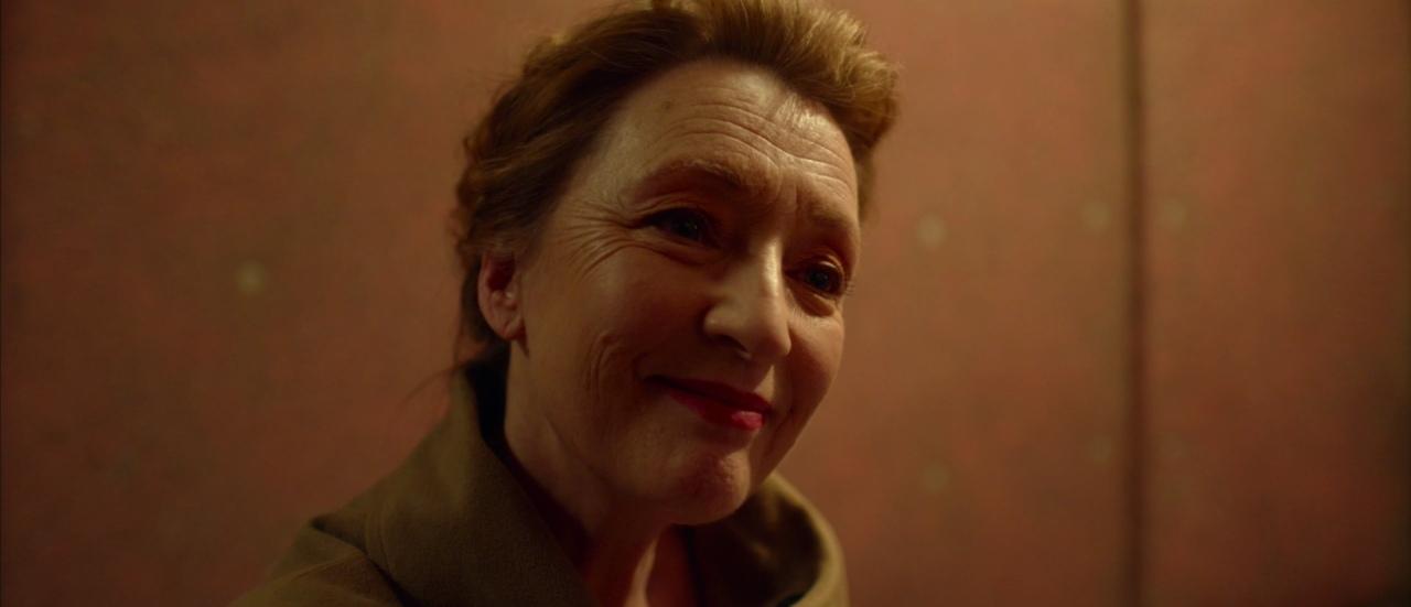 a closeup portrait of a woman with brown hair smiling in a brown coat. the woman appears to be middleaged to older and has wrinkles on her face. she is standing indoors in front of a brown background.