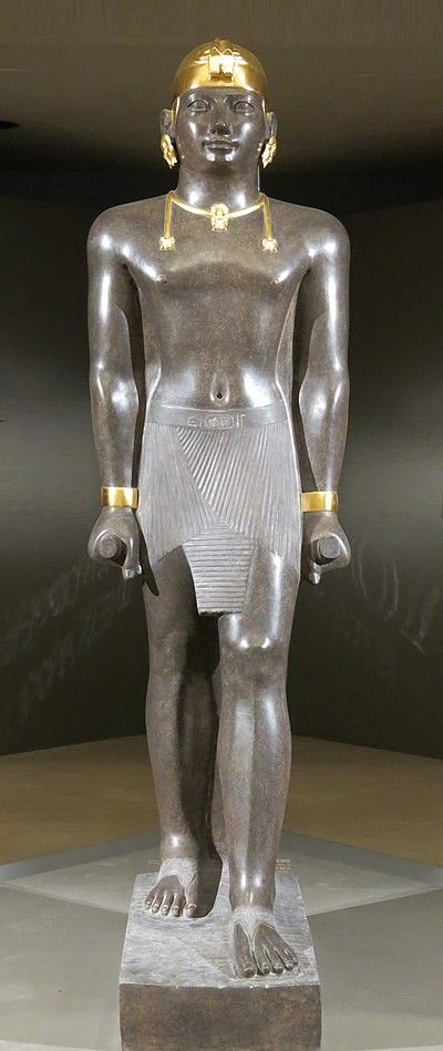 a fullshot, eyelevel view of an egyptian statue of a man with a golden headdress, necklace, bracelets, and a kilt. the statue is made of dark gray stone and is lit by studio lighting, creating a dramatic and realistic effect. the backdrop is a simple, gray wall, allowing the statue to stand out as the focal point of the image.