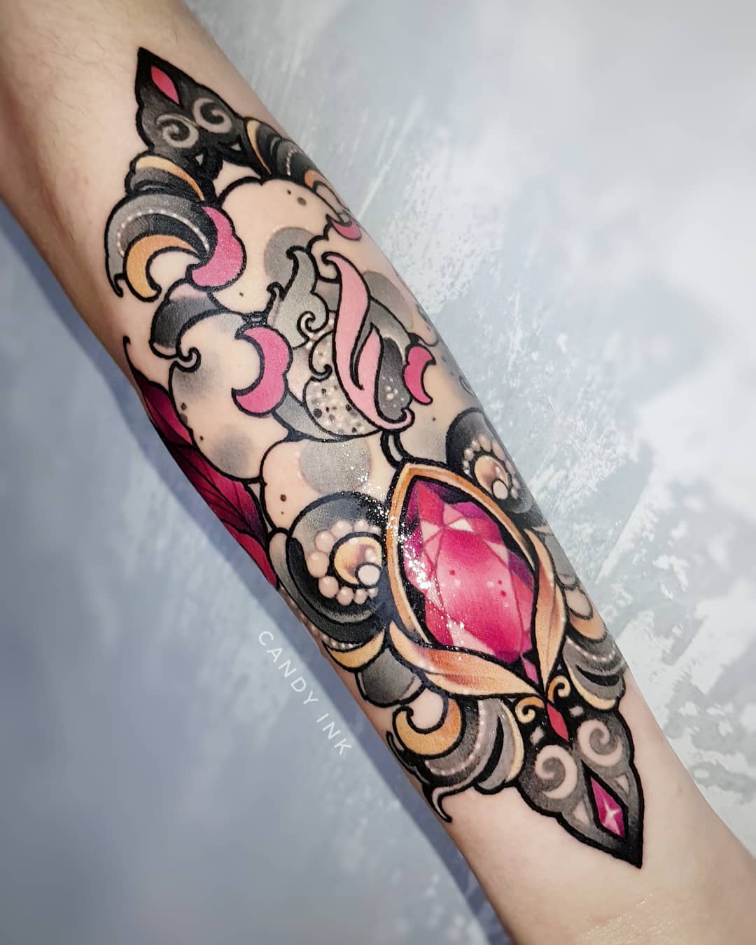 a neo traditional style tattoo of a pink and red jewel with ornate detailing on someone's forearm. the tattoo has been done in a variety of pinks, yellows, and blacks.