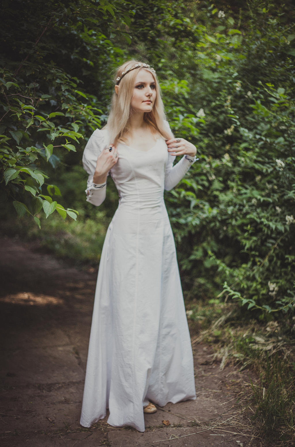 a full shot of a blonde woman wearing a long, white medievalstyle dress and a leaf crown, standing on a path in a forest. she is looking to the right, away from the camera. the forest is lush and green, and the overall atmosphere is serene and magical.  the image is styled to evoke a fairytale or fantasy setting.