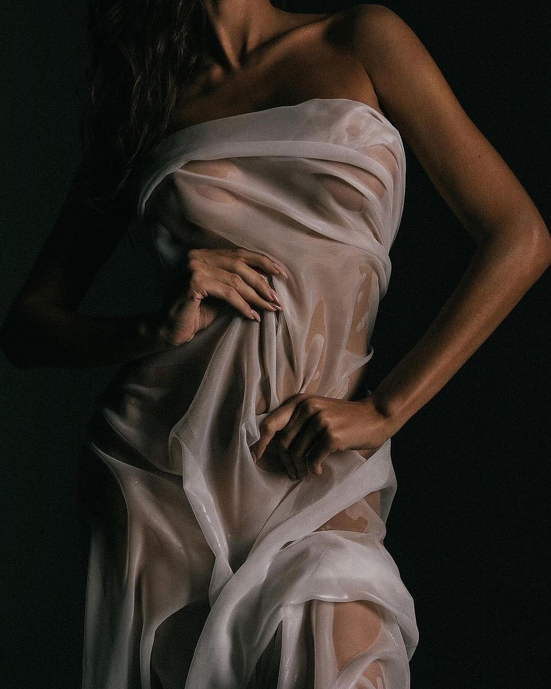 the image is a closeup studio shot of a woman's body. she is draped in a sheer, white fabric that clings to her form, revealing the contours of her body. the lighting is dramatic and moody, with deep shadows enhancing the sensual and mysterious atmosphere. the fabric appears wet or damp, adding to the overall texture and feel of the image.  the overall aesthetic is minimalist and artistic, with a focus on the interplay of light, shadow, and the woman's body.