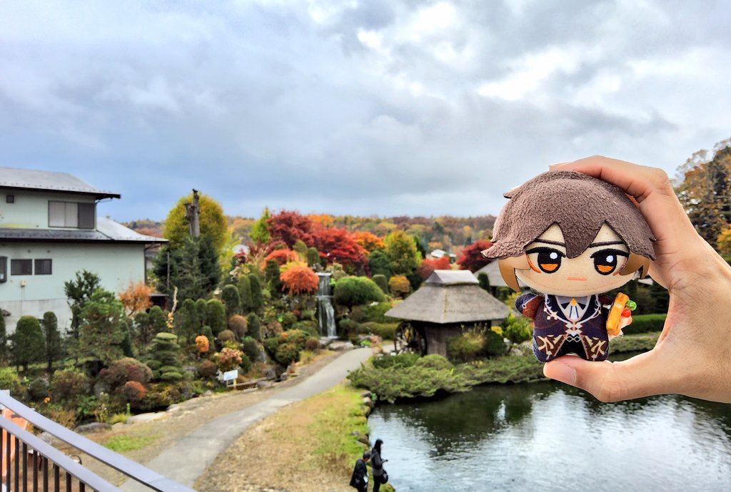 a person's hand is holding up a plush toy in front of a picturesque autumn landscape. the plush toy is an anime character with brown hair and a black suit, holding a piece of food. the background features a lush garden with a small pond, trees with vibrant foliage, and houses in the distance. the image captures the beauty of the fall season with its warm colors and natural elements.