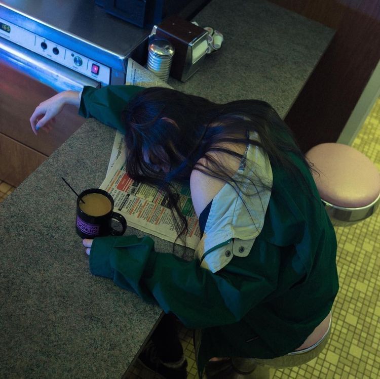 an overhead shot captures a woman slumped over a counter in a diner. she appears to be sleeping, her head resting on a newspaper. a mug of coffee sits nearby, and she's wearing a green jacket. the scene suggests fatigue or exhaustion. the diner setting includes a stool, counter, and a darkhaired woman.
