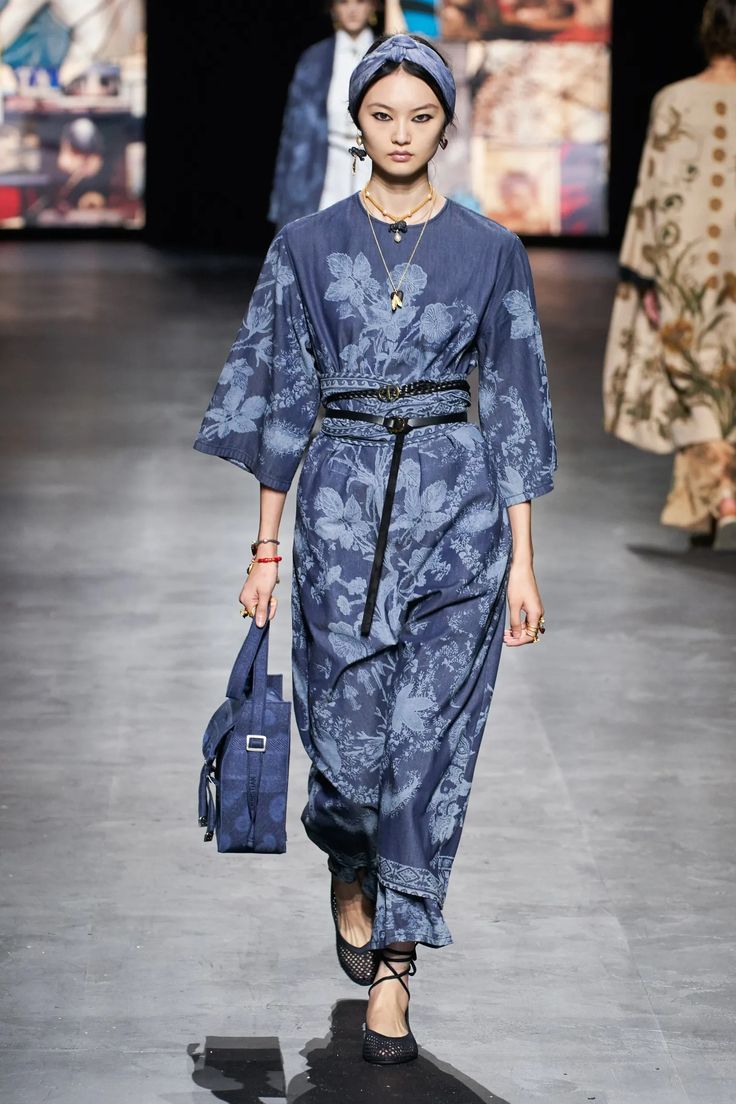 a fashion model walks the runway in a blue denim floral dress with a matching blue bag. she is wearing a black belt, black sandals and a black headband with gold jewelry. she poses for the camera as she walks down the runway.