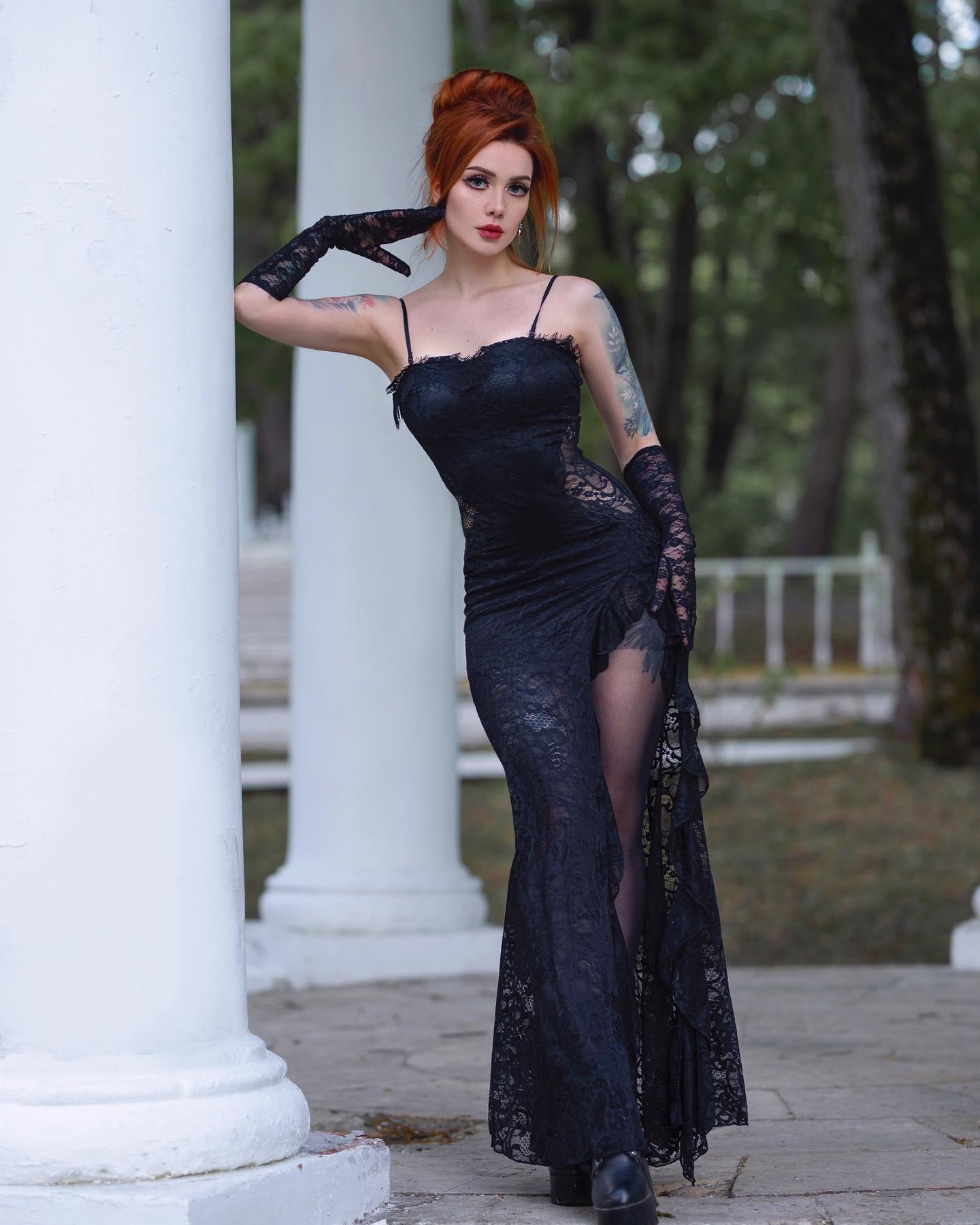 a striking redhead woman, elegantly dressed in a long black lace gown, poses between white columns in a park. her dress features a thighhigh slit and delicate lace detailing. she's accessorized with long black gloves and high heels, complementing her gothic style.  her red hair is styled up, and she has visible tattoos. the overall mood is dark, elegant, and stylish.