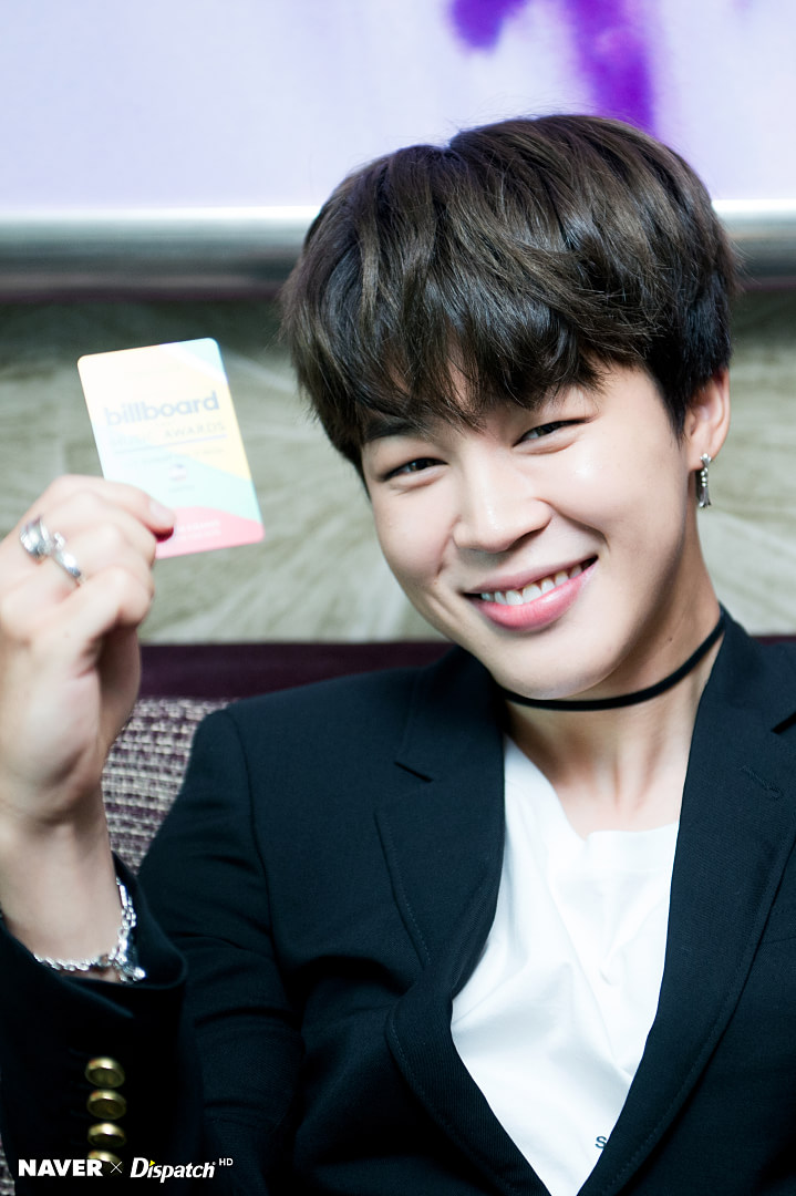 a smiling park jimin is holding a billboard award card, wearing a black suit with a white shirt and a black choker, along with a ring and bracelet.