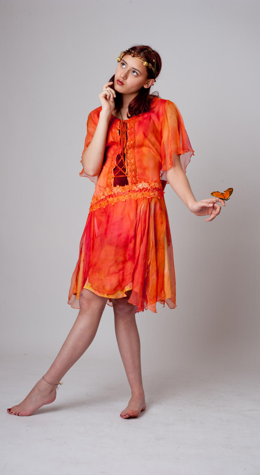a fulllength shot of a young woman wearing a flowing orange and red chiffon dress. she is barefoot and wearing a golden headband with small adornments.  a butterfly is delicately perched on her finger. she is posing in a contemplative manner, looking slightly upward,  and her overall appearance is elegant and graceful. the background is a simple light grey.