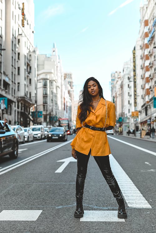 a woman stands in the middle of a city street with buildings and cars in the background. she is wearing black leggings, boots, a yellow blazer, and a black belt.