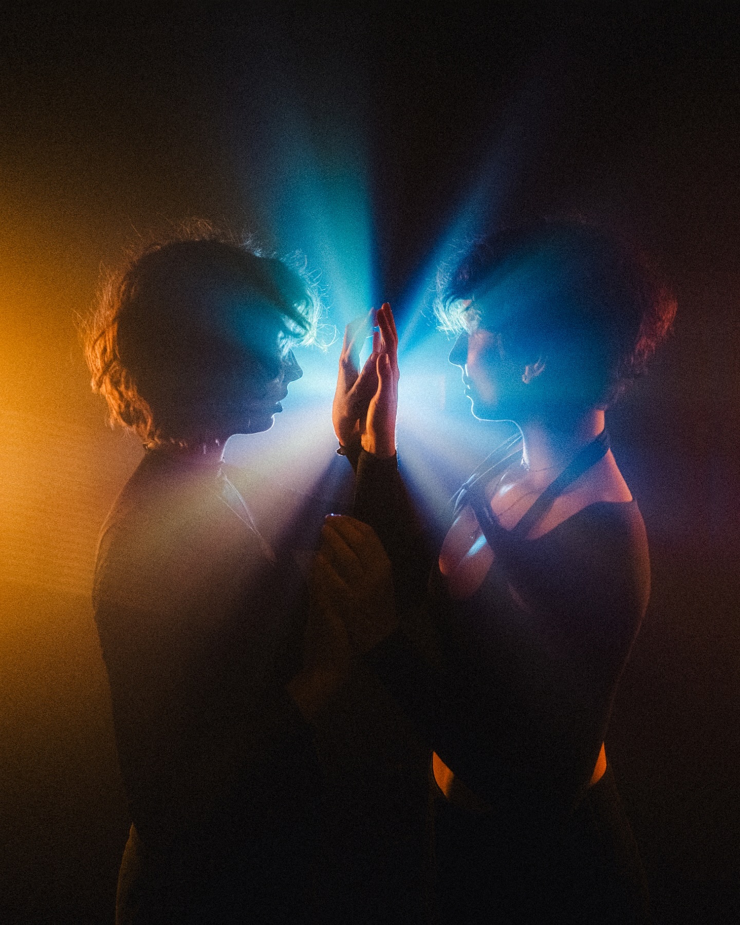 the image showcases two individuals, their silhouettes starkly defined against a dark background illuminated by dramatic blue and orange lighting.  they are embraced, their hands meeting in the center, creating a sense of intimacy and connection.  smoke or fog adds to the ethereal atmosphere. the lighting is highly stylized, with the beams seeming to emanate from between the figures, creating a mystical or spiritual ambiance. the overall mood is intense, mysterious, and artistic. the composition strongly suggests a feeling of profound connection or shared experience.