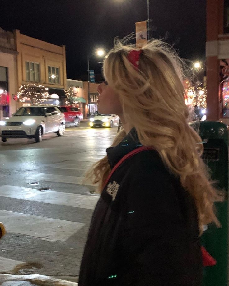 a woman with blonde hair and a red bandana is standing on a crosswalk at night in a city. there are cars driving on the street and buildings with lights in the background. she is wearing a black jacket with red straps.