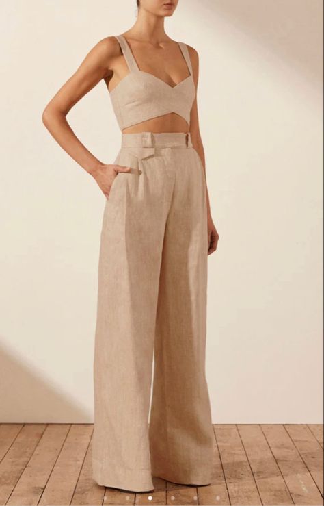 a woman is standing and wearing a beige linen crop top and matching high waisted wide leg pants. the lighting is soft and natural. the image appears to be taken in a studio setting.
