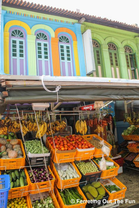 the image shows a vibrant street scene in singapore.  the focal point is a bustling fruit and vegetable stand set against the backdrop of brightly colored shophouses. the shophouses are painted in a variety of cheerful colors – blues, greens, oranges, and purples – creating a striking contrast with the fresh produce displayed in orange crates.  bananas hang from the ceiling of the stall, while various other fruits and vegetables like tomatoes, green beans, eggplants, cucumbers are neatly arranged. the overall scene is lively and indicative of a bustling local market.