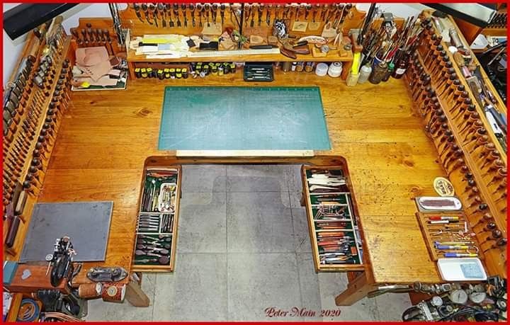 a highangle shot shows a wellorganized leatherworking workbench. the workbench is made of wood and has a ushape with a large central area. the sides are lined with various tools, primarily knives and awls, neatly arranged in holders. drawers on both sides are open, revealing more tools. a green cutting mat is placed in the center of the main working area. a shelf above holds additional supplies, including paint bottles, jars, and leather pieces. on the left side of the bench, a vintage motorcycle model sits next to other tools and supplies. pens, markers, and a ruler are on the right side. the image highlights the organized and functional layout of the workspace.