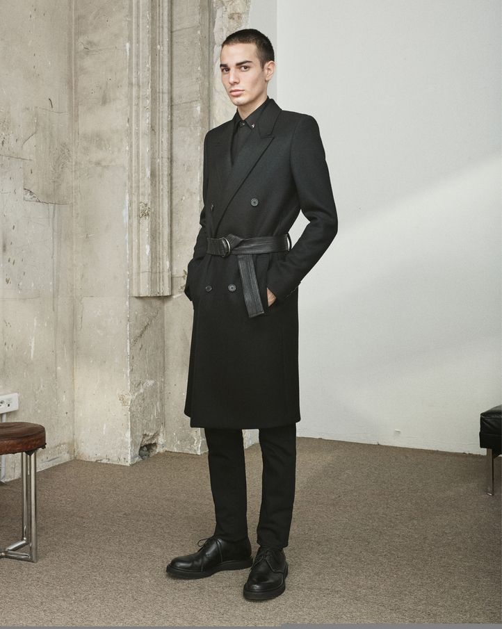 a man stands indoors wearing a black doublebreasted overcoat cinched at the waist with a black belt. he is also wearing black pants and black shoes. the background is a textured wall.