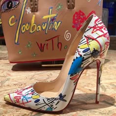 a white pair of high heels with red soles with a graffitistyle design, sitting on a brown  bag with the word "louboutin" and "with" written on it.  the shoes are a statement piece with a very fashionable and eyecatching design.