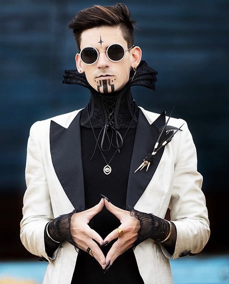 a man dressed in a white tuxedo with a black satin lapel, a black lace choker and black gloves with feather embellishments. he has a black star tattooed on his forehead and a black stripe design on his neck. he is wearing dark round sunglasses and has long, black nail polish on his fingers. he stands with his hands clasped in front of him, looking intense. he is looking directly at the camera, with a serious expression on his face. the background is a dark blue wall.