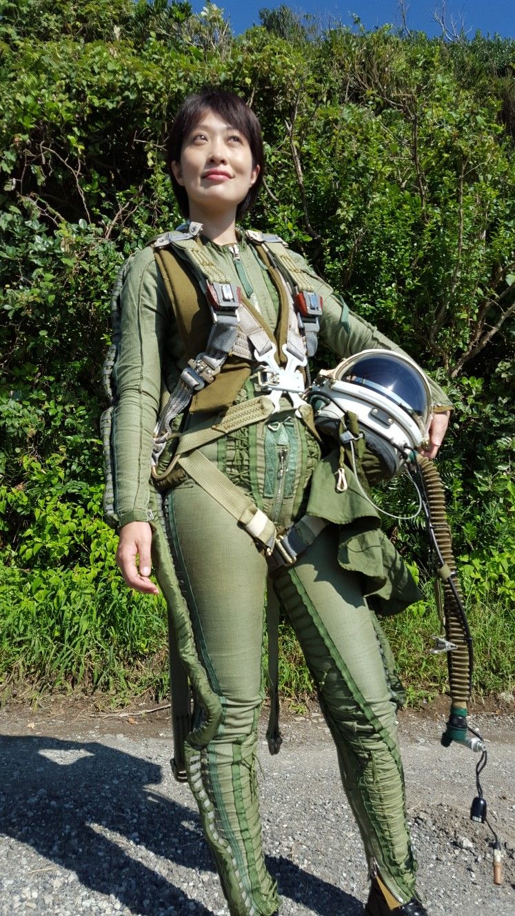 a woman in a green jumpsuit and a helmet looks up at the sky, standing in a forest with a dirt path in front of her. she appears to be a pilot, possibly an astronaut.