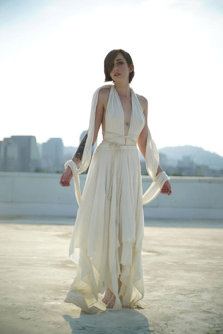 a woman in a long, flowing offwhite dress stands barefoot on a rooftop, against a backdrop of a city skyline. the dress is simple yet elegant, with a halter neck and layered fabric, creating a bohemian feel. the woman has short, dark hair and a calm, serene expression. the overall image is bright and airy, with the light reflecting off the fabric of the dress.